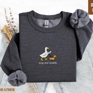 May include: Dark heather gray sweatshirt with a cartoon duck walking a dog on a leash. The text "wag and waddle" is below the graphic. The sweatshirt has a tag that says "Gildan" and the words "Super Soft" are in a brown box.