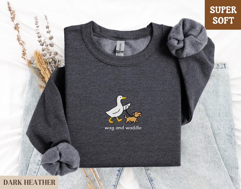 May include: Dark heather gray sweatshirt with a cartoon duck walking a dog on a leash. The text "wag and waddle" is below the graphic. The sweatshirt has a tag that says "Gildan" and the words "Super Soft" are in a brown box.