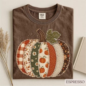 May include: A dark brown t-shirt featuring a large, central pumpkin graphic composed of various fabric-style patches in plaid, floral, and herringbone patterns. The pumpkin design includes small orange pumpkin illustrations and a green leaf.