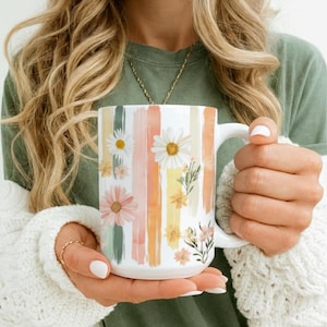 Puede incluir: Taza de cerámica blanca con diseño floral. La taza presenta rayas verticales en colores pastel, incluyendo rosa, naranja y verde, adornadas con ilustraciones de margaritas y flores silvestres. La taza tiene un asa grande.