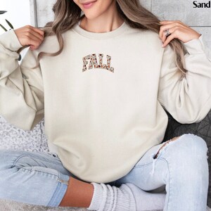 May include: A beige sweatshirt with a brown and white patterned "FALL" graphic on the front.