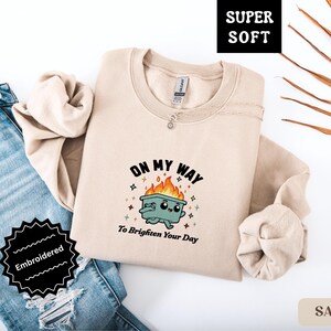 Pu&ograve; includere: Felpa color sabbia con la scritta "ON MY WAY To Brighten Your Day" e un bidone della spazzatura dei cartoni animati in fiamme. La felpa &egrave; ricamata ed etichettata "Super Soft".
