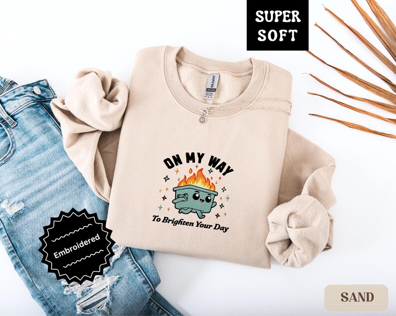 Pu&ograve; includere: Felpa color sabbia con la scritta "ON MY WAY To Brighten Your Day" e un bidone della spazzatura dei cartoni animati in fiamme. La felpa &egrave; ricamata ed etichettata "Super Soft".