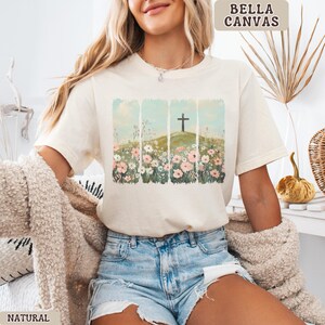 May include: Cream-colored t-shirt with a graphic of a cross on a hill, surrounded by flowers. The design is set against a backdrop of blue sky and vertical stripes. The shirt is labeled "BELLA CANVAS" and "NATURAL".