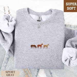K&ouml;nnte beinhalten: Hellgraues Sweatshirt mit drei gestickten Pferden in Braun- und Beiget&ouml;nen. Das Sweatshirt hat einen Rundhalsausschnitt und lange &Auml;rmel. Die Worte "SUPER SOFT" stehen in einer braunen Box und "SPORT GRAY" in einer beigen Box.