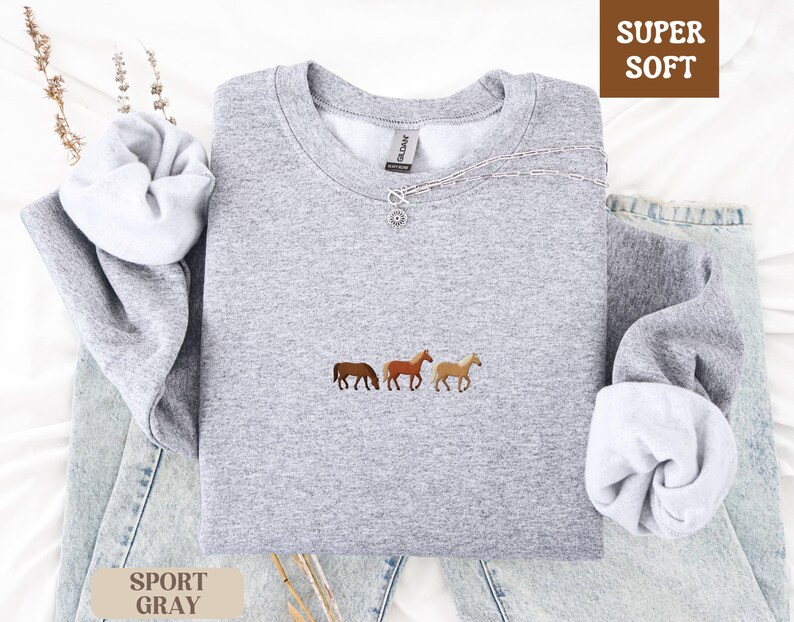 K&ouml;nnte beinhalten: Hellgraues Sweatshirt mit drei gestickten Pferden in Braun- und Beiget&ouml;nen. Das Sweatshirt hat einen Rundhalsausschnitt und lange &Auml;rmel. Die Worte "SUPER SOFT" stehen in einer braunen Box und "SPORT GRAY" in einer beigen Box.