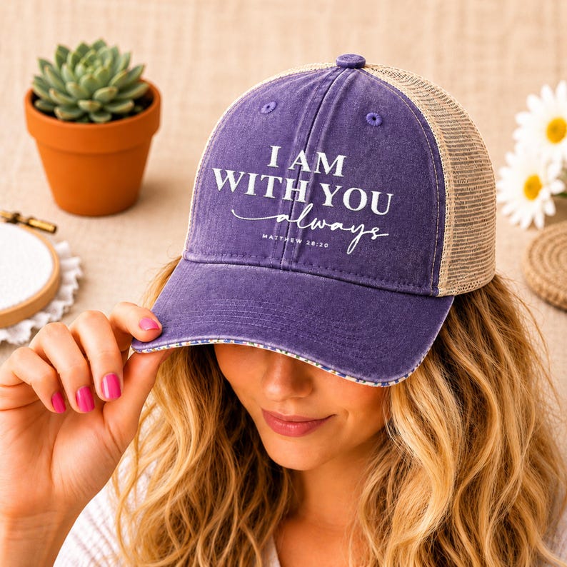 May include: A purple and tan trucker hat with the text "I AM WITH YOU always" in white script. The hat has a curved brim and a mesh back. The hat is a casual accessory.