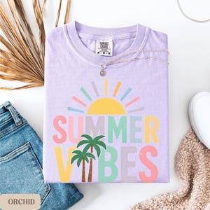 K&ouml;nnte beinhalten: Ein fliederfarbenes Comfort Colors T-Shirt mit dem Schriftzug "SUMMER VIBES" in bunten Buchstaben und einer Sonne und Palmen-Grafik. Das gefaltete Shirt wird mit einer silbernen Halskette, Jeans und einem wei&szlig;en Schuh pr&auml;sentiert.