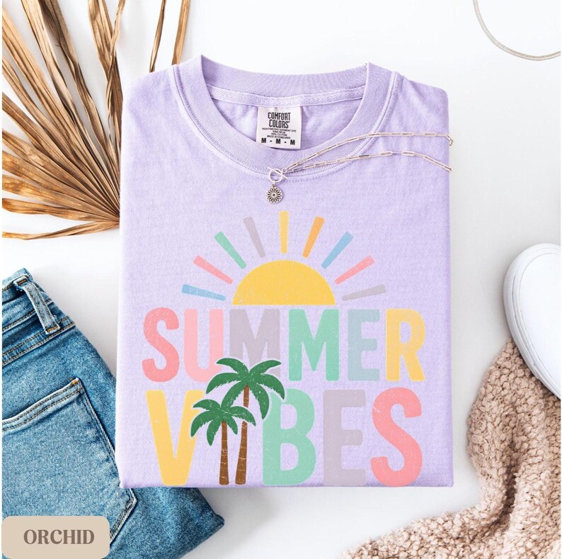 K&ouml;nnte beinhalten: Ein fliederfarbenes Comfort Colors T-Shirt mit dem Schriftzug "SUMMER VIBES" in bunten Buchstaben und einer Sonne und Palmen-Grafik. Das gefaltete Shirt wird mit einer silbernen Halskette, Jeans und einem wei&szlig;en Schuh pr&auml;sentiert.