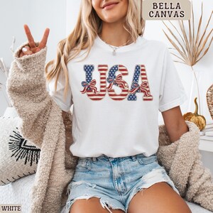May include: White t-shirt with the word "USA" in red, white, and blue, with a patriotic bow design. The shirt is made of a soft material and is perfect for celebrating Independence Day. The brand name "BELLA CANVAS" is printed on the top right.