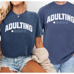 May include: Two navy blue t-shirts with the word "ADULTING" in white, arched text above five stars. The shirts are a comfortable fit, and the image includes the words "COMFORT COLORS" and "DENIM".