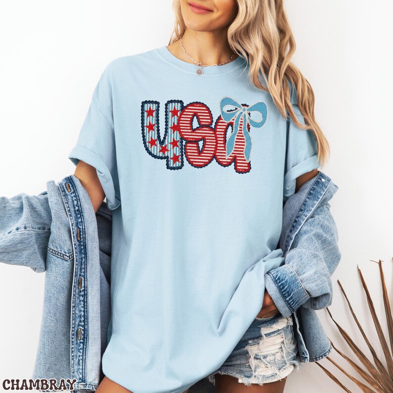 Op de afbeelding: Lichtblauw T-shirt met het woord "USA" in rood, wit en blauw. De letters "U" en "A" zijn versierd met rode sterren en strepen, en de "S" heeft rode en witte strepen. Aan de rechterkant zit een blauwe strik.