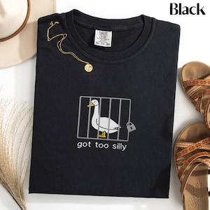 May include: Black crew neck t-shirt with an embroidered design of a duck in a cage, the text "got too silly" is below the duck. A gold necklace and brown sandals are also in the image.