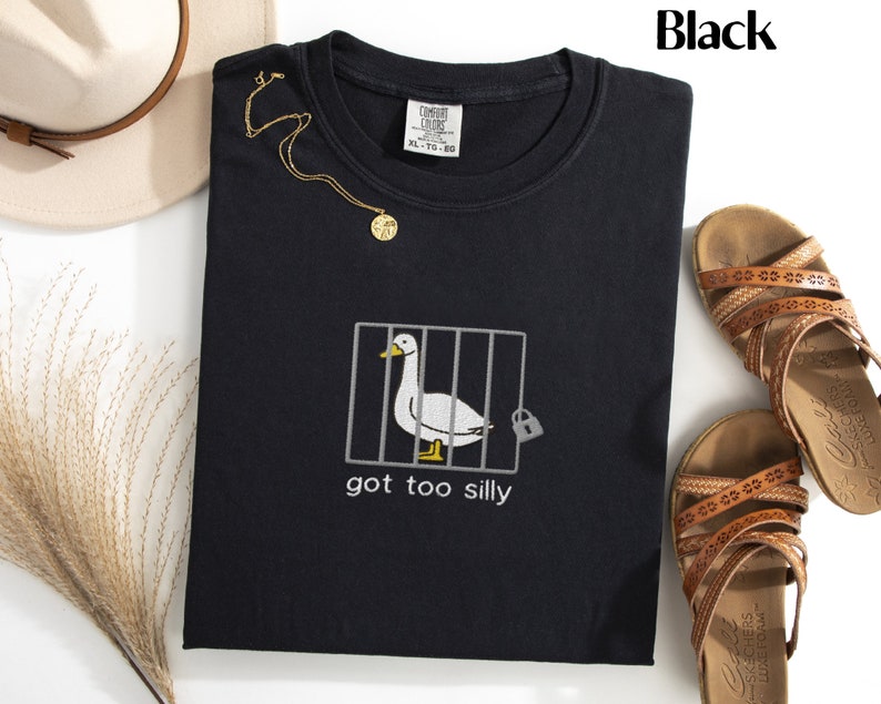 May include: Black crew neck t-shirt with an embroidered design of a duck in a cage, the text "got too silly" is below the duck. A gold necklace and brown sandals are also in the image.