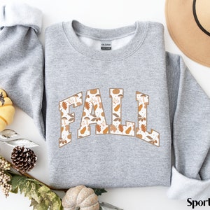 K&ouml;nnte beinhalten: Ein grauer Pullover mit dem Wort "FALL" in Wei&szlig; bedruckt, mit einem braunen und orangefarbenen Bl&auml;ttermuster. Der Pullover ist ein Rundhals-Modell und hat eine lockere Passform.