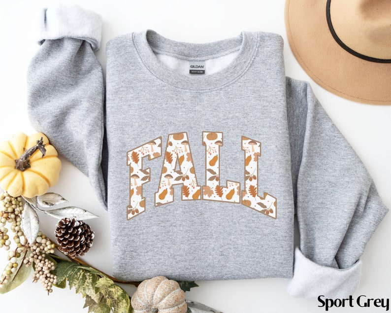 K&ouml;nnte beinhalten: Ein grauer Pullover mit dem Wort "FALL" in Wei&szlig; bedruckt, mit einem braunen und orangefarbenen Bl&auml;ttermuster. Der Pullover ist ein Rundhals-Modell und hat eine lockere Passform.