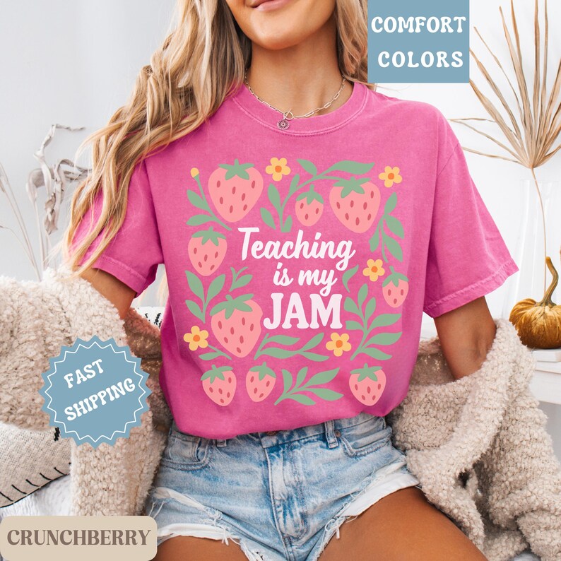 May include: A pink t-shirt with the text "Teaching is my JAM" surrounded by a strawberry and floral design. The shirt has short sleeves and a crew neck. The words "Comfort Colors" are in the upper right corner.