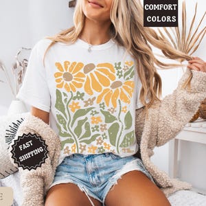May include: A white t-shirt featuring a graphic print of yellow sunflowers and green foliage with a distressed texture. The shirt is paired with blue denim shorts and a beige textured cardigan. Text on the image includes COMFORT COLORS.