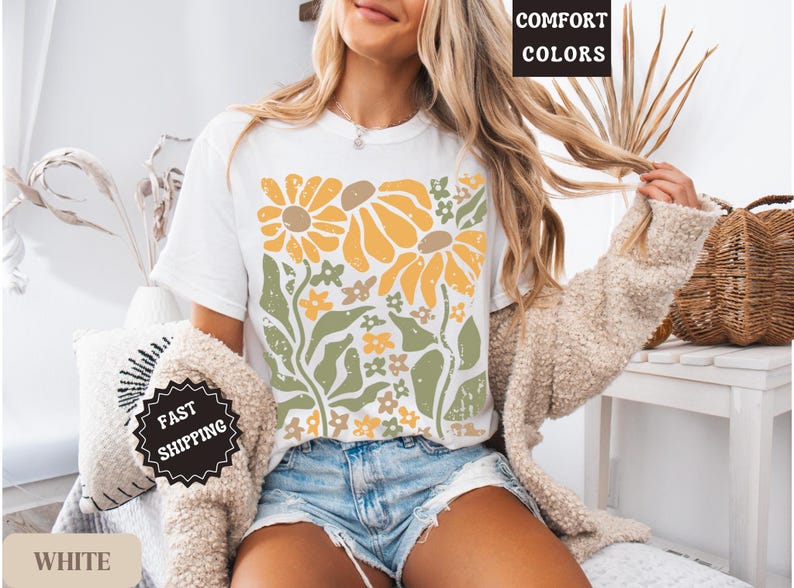 May include: A white t-shirt featuring a graphic print of yellow sunflowers and green foliage with a distressed texture. The shirt is paired with blue denim shorts and a beige textured cardigan. Text on the image includes COMFORT COLORS.