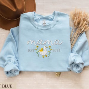 May include: Light blue sweatshirt with the text "mama est 2025" and an embroidered image of a duck with a baby duck.