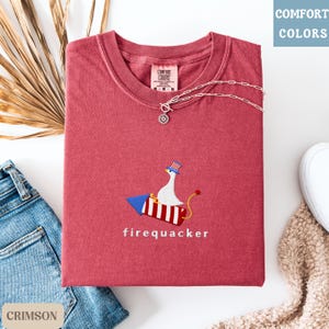 K&ouml;nnte beinhalten: Karmesinrotes T-Shirt mit einem skurrilen gestickten Design einer Gans auf einer Rakete, mit der Aufschrift "firequacker". Das Shirt hat einen Rundhalsausschnitt und kurze &Auml;rmel. Eine silberne Halskette mit Anh&auml;nger liegt &uuml;ber dem Shirt.