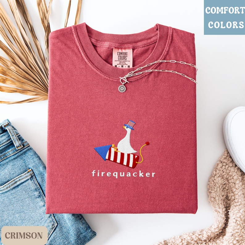 K&ouml;nnte beinhalten: Karmesinrotes T-Shirt mit einem skurrilen gestickten Design einer Gans auf einer Rakete, mit der Aufschrift "firequacker". Das Shirt hat einen Rundhalsausschnitt und kurze &Auml;rmel. Eine silberne Halskette mit Anh&auml;nger liegt &uuml;ber dem Shirt.
