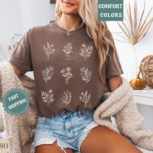 May include: A brown t-shirt with a white floral design. The shirt has the text "Comfort Colors" printed on the top left corner and "Fast Shipping" printed on the bottom left corner.
