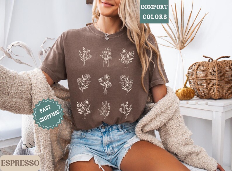 May include: A brown t-shirt with a white floral design. The shirt has the text "Comfort Colors" printed on the top left corner and "Fast Shipping" printed on the bottom left corner.