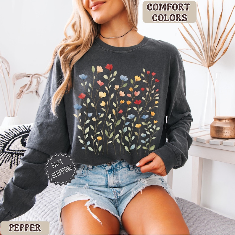 May include: Dark gray long-sleeve sweatshirt with a floral design. The graphic features colorful flowers in red, blue, yellow, and orange. The words "COMFORT COLORS" are printed at the top. The word "PEPPER" is printed at the bottom.