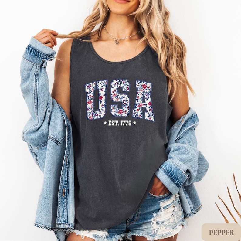 K&ouml;nnte beinhalten: Dunkelgraues Tanktop mit dem Wort "USA" in einem floralen Muster. Darunter steht "EST. 1776". Das Tanktop wird mit einer Jeansjacke und Jeansshorts kombiniert.