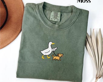 May include: Moss green t-shirt with a crew neck. The shirt features an embroidered design of a white duck walking a small brown dog on a leash. The tag reads "Comfort Colors".