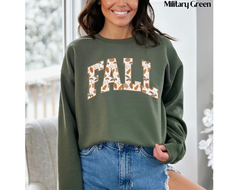May include: A military green sweatshirt with the word "FALL" in a white and brown floral pattern. The sweatshirt has a crew neck and long sleeves. The model is wearing blue denim shorts.