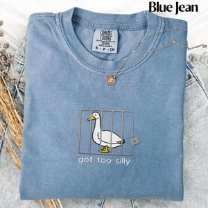 May include: A blue Comfort Colors t-shirt with an embroidered design of a white duck behind bars, with the text "got too silly" below. A silver necklace with a pendant is draped over the shirt. The words "Blue Jean" are at the top.