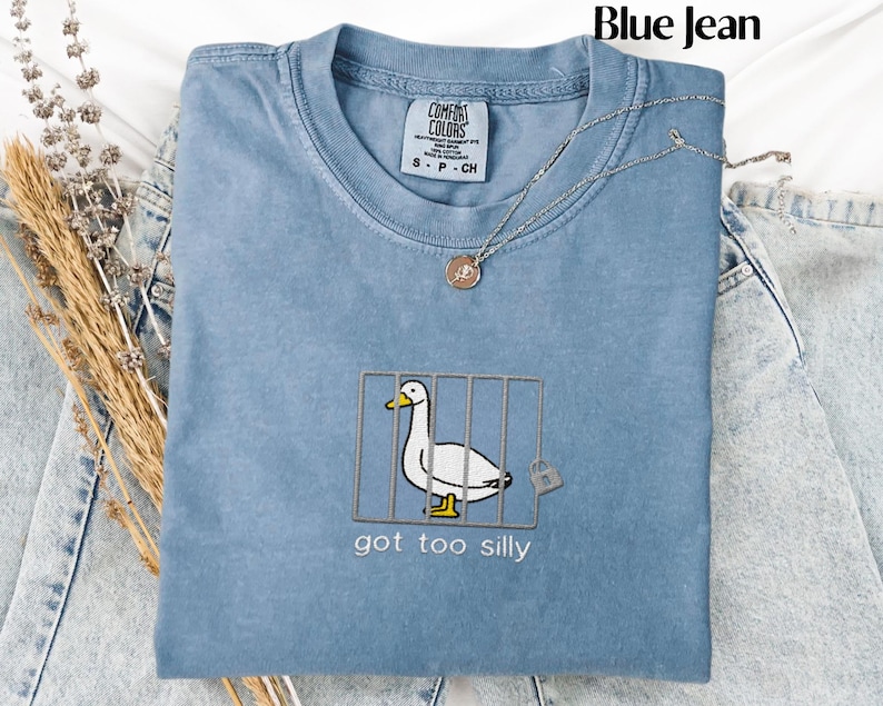 May include: A blue Comfort Colors t-shirt with an embroidered design of a white duck behind bars, with the text "got too silly" below. A silver necklace with a pendant is draped over the shirt. The words "Blue Jean" are at the top.