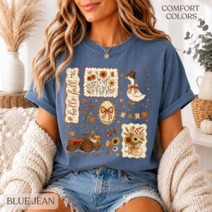 May include: A blue short-sleeve t-shirt featuring a graphic print with the text hello fall, a tractor, a goose wearing a hat, sunflowers, and autumn foliage. The design includes various rectangular and oval frames with plaid and floral patterns.