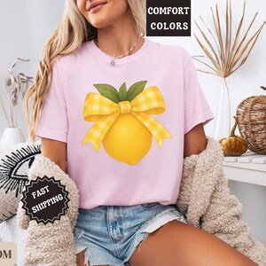 Puede incluir: Una camiseta rosa con un lim&oacute;n amarillo con un lazo de cuadros amarillos y blancos en la parte superior. El texto "COMFORT COLORS" est&aacute; en una etiqueta blanca unida a la camiseta.