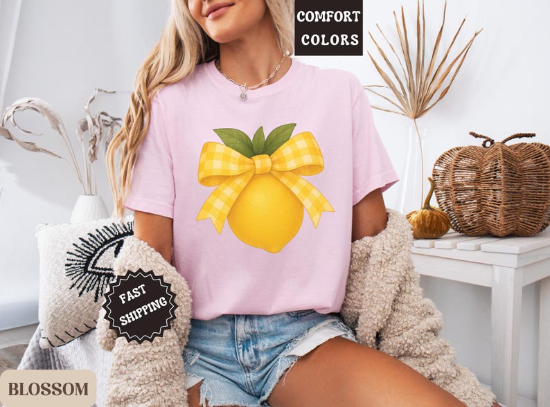 Puede incluir: Una camiseta rosa con un lim&oacute;n amarillo con un lazo de cuadros amarillos y blancos en la parte superior. El texto "COMFORT COLORS" est&aacute; en una etiqueta blanca unida a la camiseta.