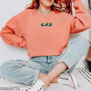 May include: Terracotta-colored sweatshirt with a crew neck and long sleeves. The sweatshirt features a small green embroidered design of three alligators. The word "TERRACOTTA" is printed in a serif font at the bottom of the image. The model is wearing light blue jeans and white sneakers.