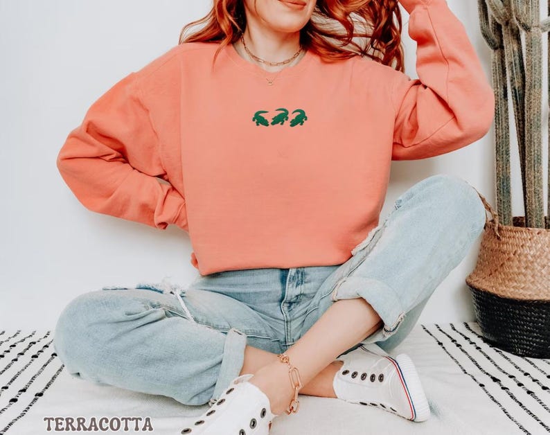 May include: Terracotta-colored sweatshirt with a crew neck and long sleeves. The sweatshirt features a small green embroidered design of three alligators. The word "TERRACOTTA" is printed in a serif font at the bottom of the image. The model is wearing light blue jeans and white sneakers.
