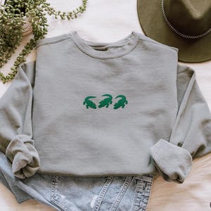 May include: A gray sweatshirt with three green alligator designs embroidered on the front. The long-sleeved crew neck shirt is folded on top of blue jeans. A green hat and a potted plant are in the background.