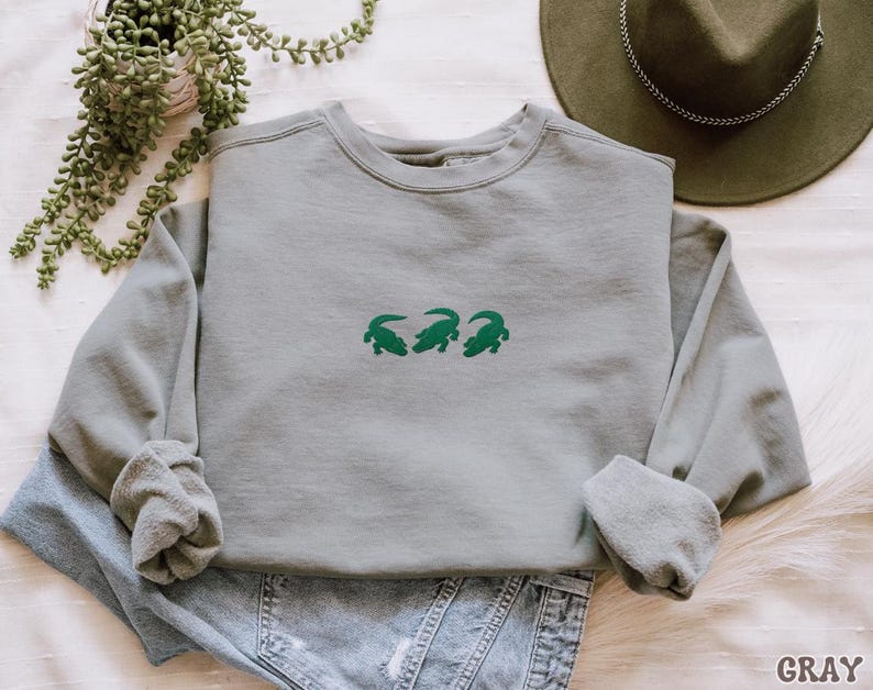 May include: A gray sweatshirt with three green alligator designs embroidered on the front. The long-sleeved crew neck shirt is folded on top of blue jeans. A green hat and a potted plant are in the background.