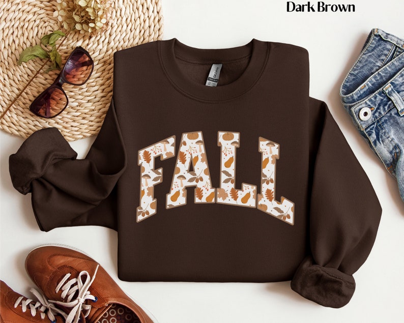 May include: Dark brown sweatshirt with the word "FALL" in large, block letters. The letters are filled with a pattern of fall leaves, mushrooms, and pumpkins in shades of brown, beige, and orange. The sweatshirt has a crew neck and long sleeves.