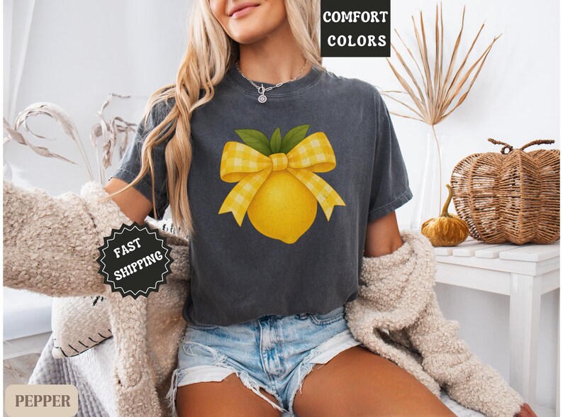 Puede incluir: Una camiseta gris oscuro con un lim&oacute;n amarillo y un lazo de cuadros. El texto "COMFORT COLORS" es visible en el fondo.