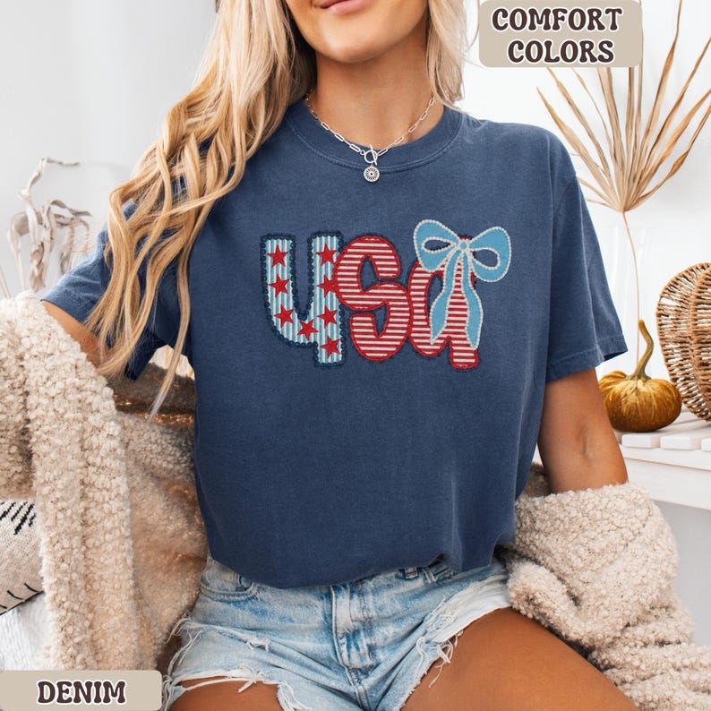 Op de afbeelding: Een marineblauw t-shirt met het woord "USA" geborduurd in rood, wit en blauw. De "U" heeft rode sterren op een blauwe achtergrond, de "S" heeft rood-witte strepen en de "A" is versierd met een blauwe strik. Het shirt heeft de tekst "Comfort Colors" en "Denim".