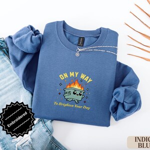 Pu&ograve; includere: Felpa blu indaco con la frase ricamata "ON MY WAY To Brighten Your Day" e una grafica di un bidone della spazzatura in fiamme. La felpa &egrave; abbinata a jeans blu e una collana d'argento.