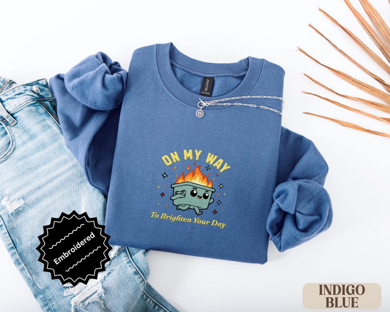 Pu&ograve; includere: Felpa blu indaco con la frase ricamata "ON MY WAY To Brighten Your Day" e una grafica di un bidone della spazzatura in fiamme. La felpa &egrave; abbinata a jeans blu e una collana d'argento.