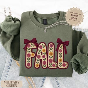 May include: A military green sweatshirt featuring the word "FALL" in a floral design. The letters are beige with burgundy outlines and bows, filled with pink and yellow flowers. The sweatshirt has a crew neck and long sleeves.
