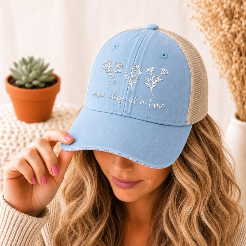 May include: A light blue baseball cap with a mesh back and a curved brim. The cap features embroidered floral designs and the phrase "one day at a time" in silver lettering. The cap is worn by a person.