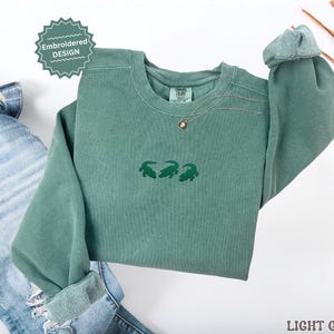 May include: A light green sweatshirt with three embroidered green alligators. The sweatshirt has a crew neck and long sleeves. The words "Embroidered Design" are on a green sticker. The words "Light Green" are at the bottom right.