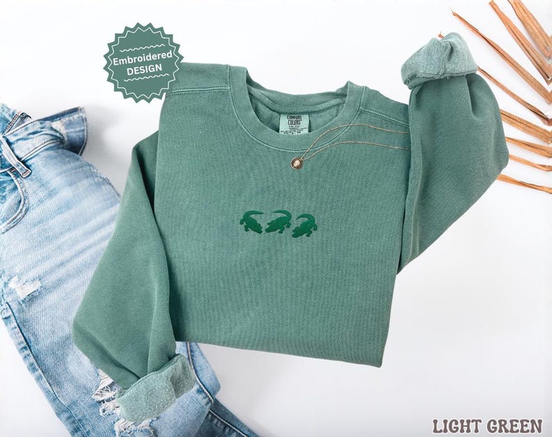 May include: A light green sweatshirt with three embroidered green alligators. The sweatshirt has a crew neck and long sleeves. The words "Embroidered Design" are on a green sticker. The words "Light Green" are at the bottom right.